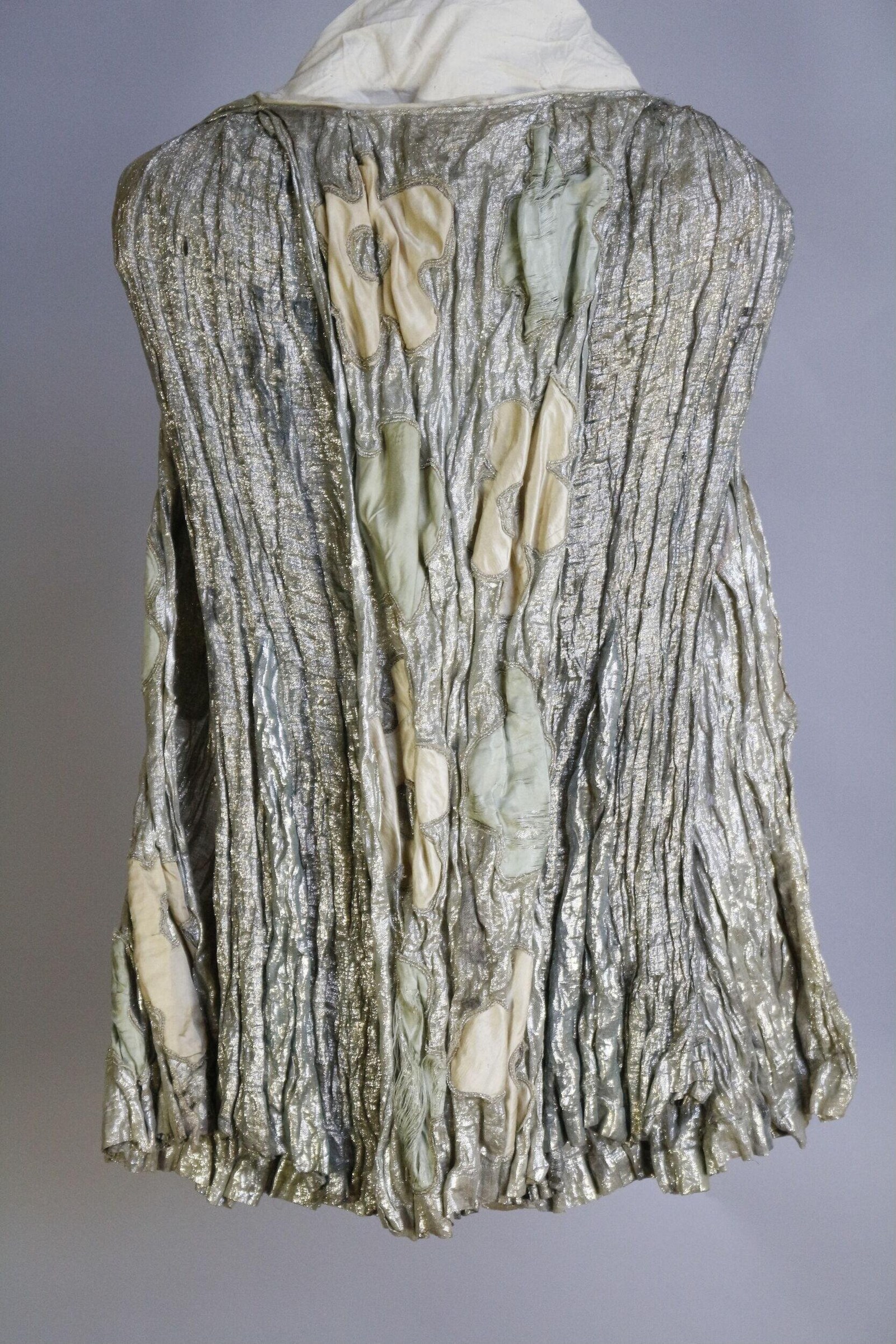 Back view of a sleeveless theatrical costume in pleated metallic silver fabric with cream and pale green appliqué shapes, gathered hem, and white neckline.