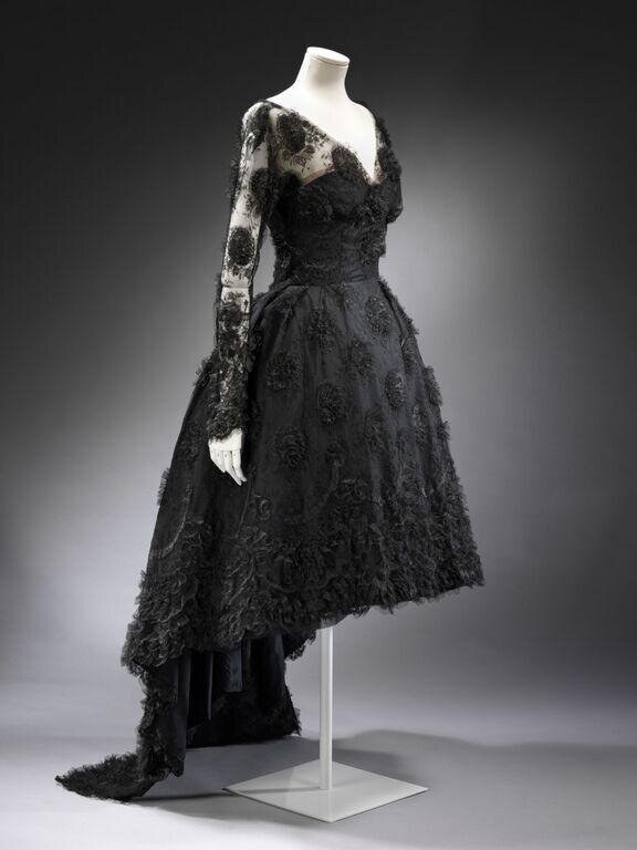 Black lace evening dress with asymmetrical hemline on mannequin, displayed from three-quarter angle with extended train.