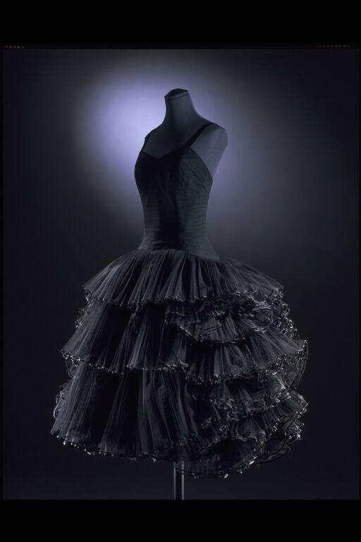 Headless dress mannequin wearing a fitted dark bodice and dramatic tiered tulle skirt with beaded edges, backlit against dark background.