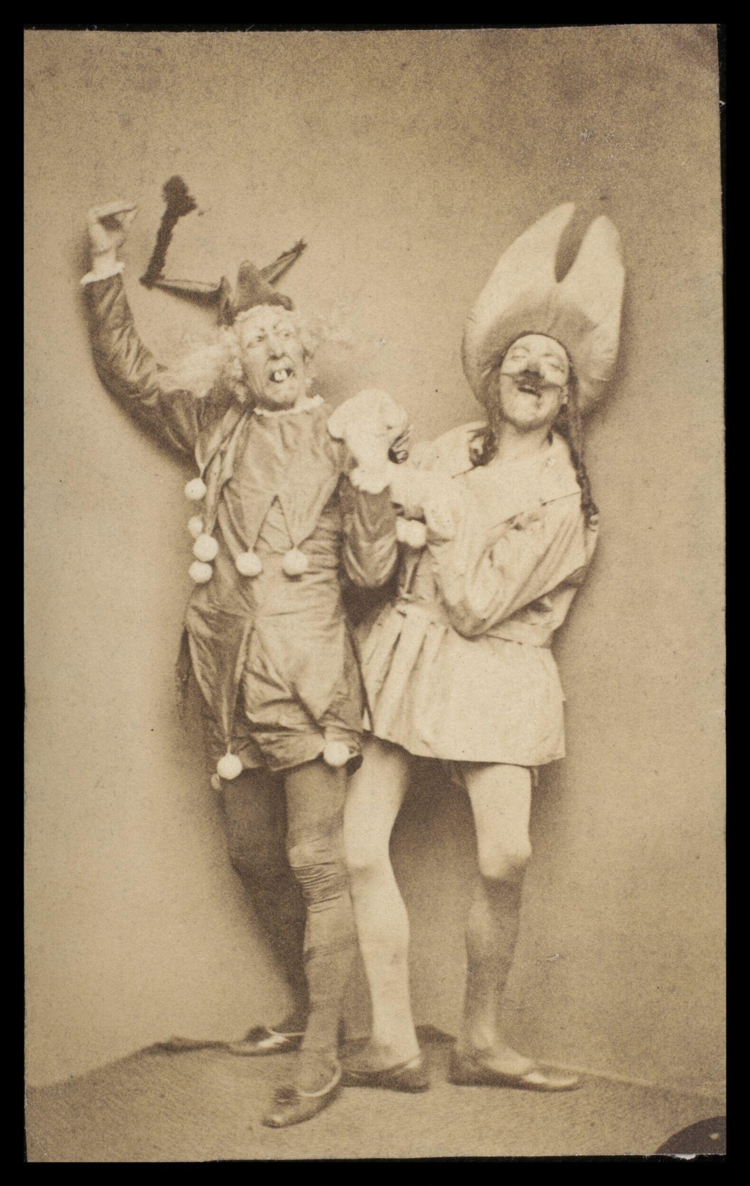 Two Victorian theatrical performers in elaborate costumes and tall headdresses grin at the camera, striking playful poses in a studio setting.