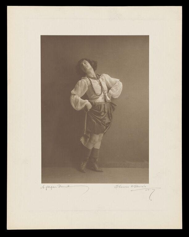 Woman in white blouse and rust skirt stands with head tilted back and arms positioned expressively against neutral backdrop