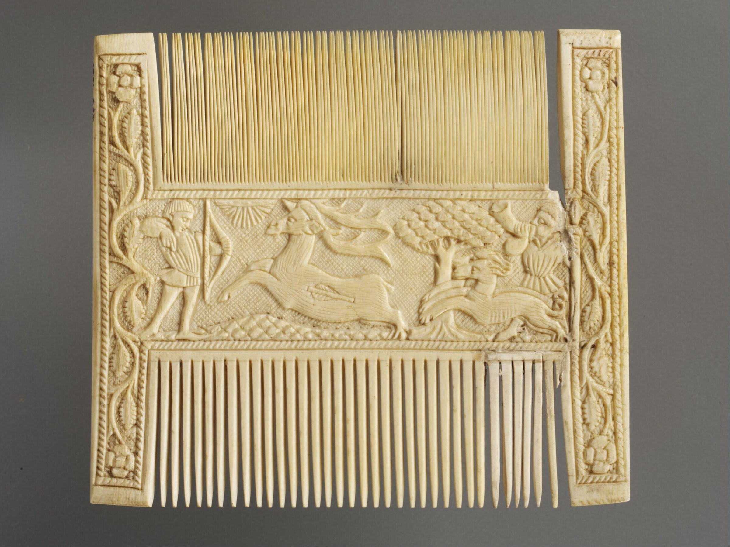 Carved ivory comb showing hunting above and dancing figures below in 15th-century style, with fine relief carving and decorative borders.