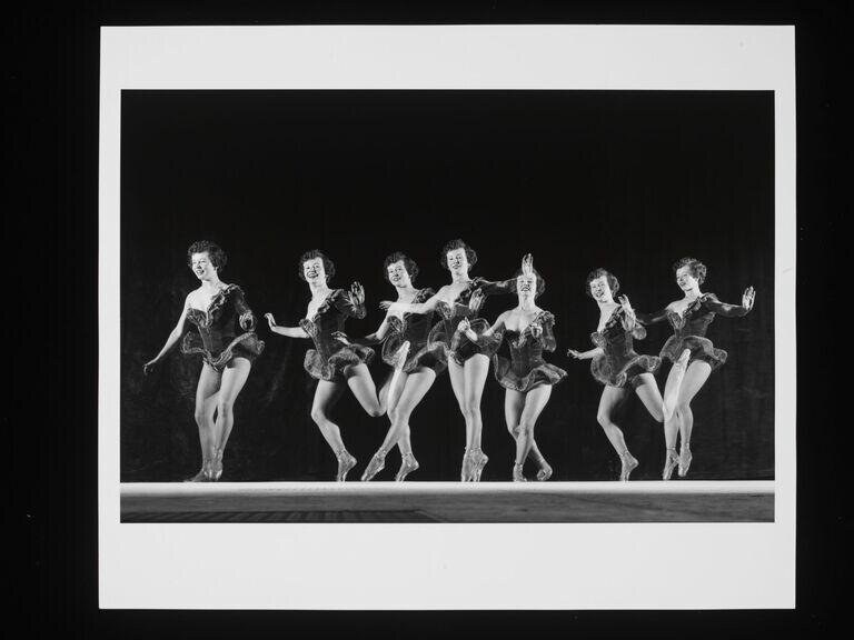 Seven female dancers in identical sequined costumes stand in a line on a dark stage, arms raised in synchronized gesture, evenly lit from the front.