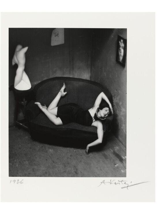 A woman in a dark dress lies twisted in an armchair, one leg raised, arms angular, in an exaggerated contorted pose.