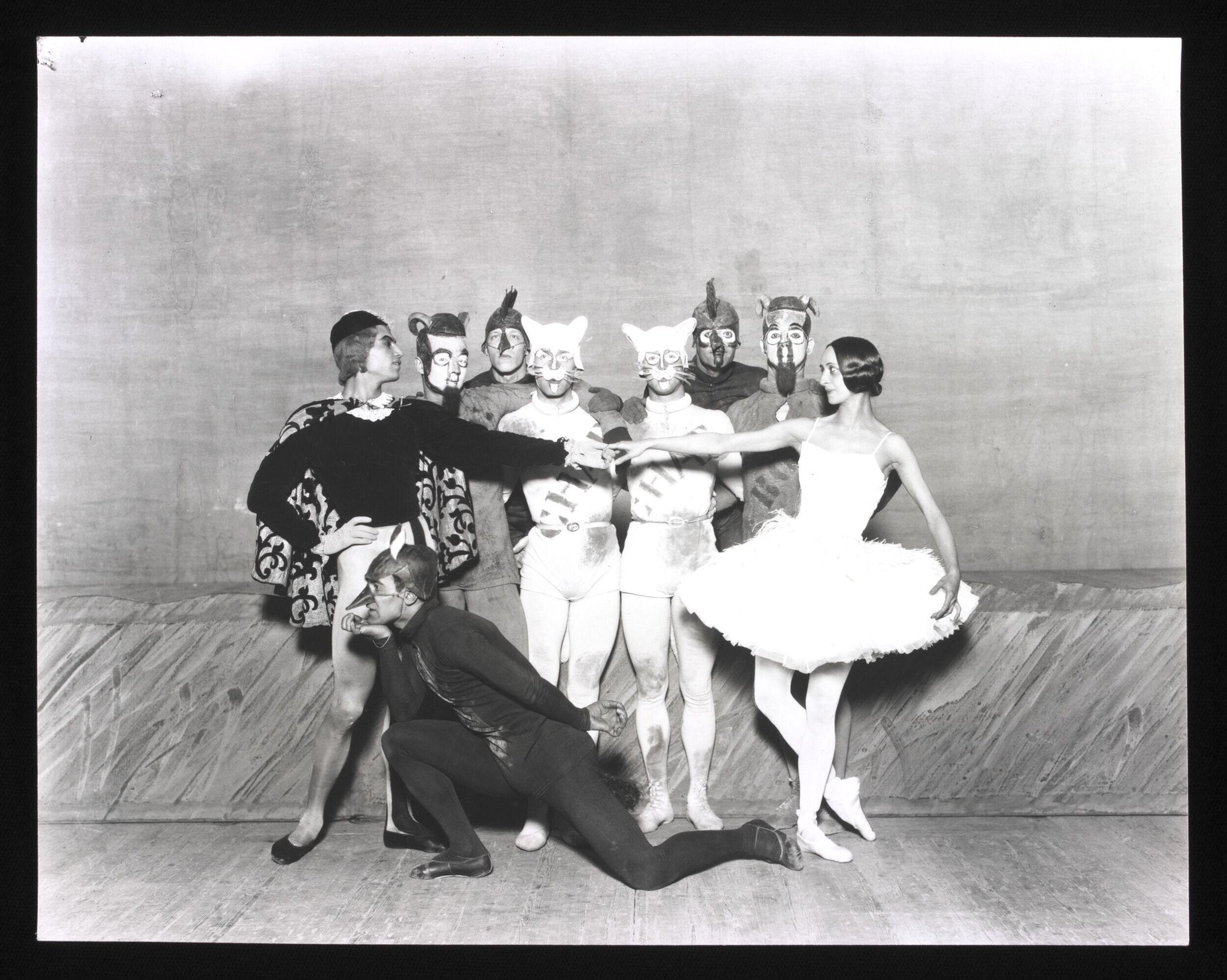 Eight costumed dancers: seven wearing animal masks stand on a wooden stage while an unmasked man in ornate dress kneels toward them.