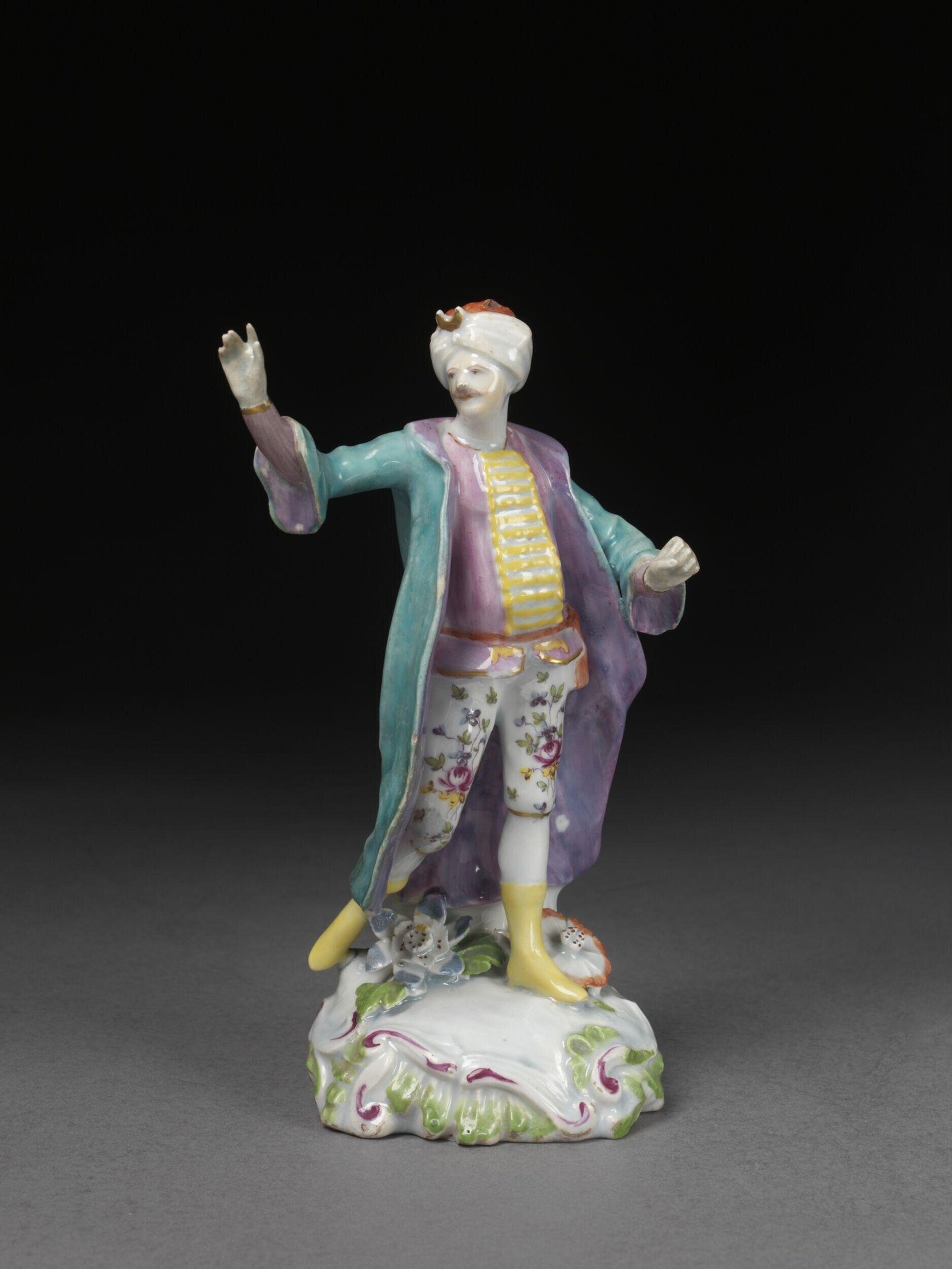 Porcelain figure of a man in white turban and purple-and-teal Ottoman-style coat, gesturing with raised right arm on a decorated rococo base.