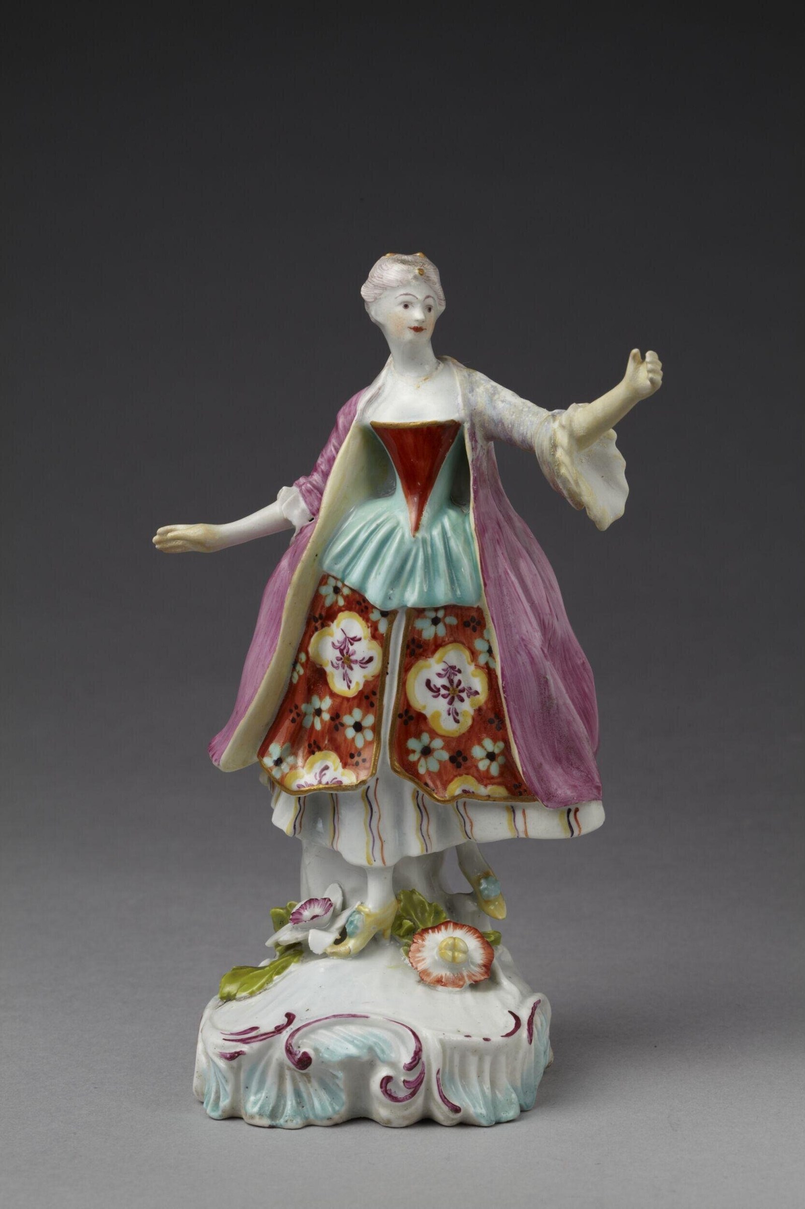 Porcelain figurine of a woman in layered red and blue dress with pink wrap, arms extended in greeting, on a rococo base.