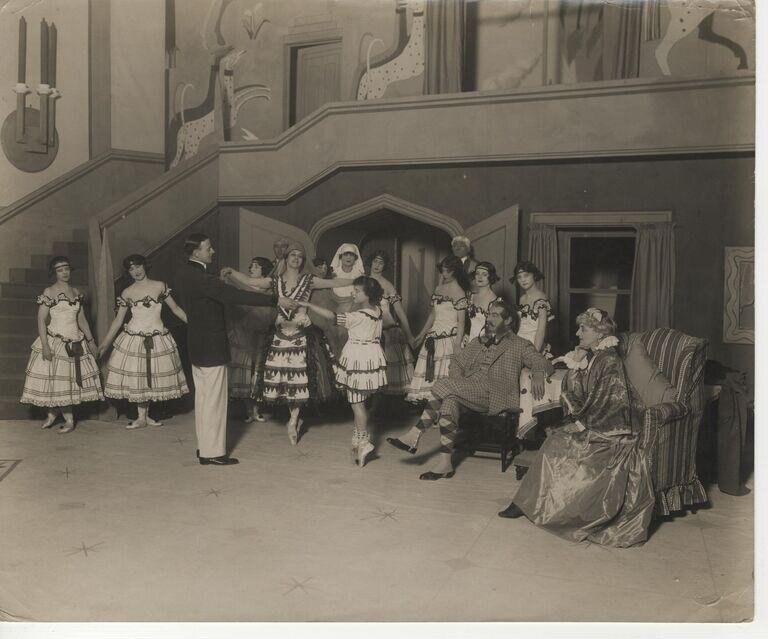A studio photograph of a dozen costumed performers posed stiffly in a stylized Art Deco interior, centered on an ornately dressed woman in an elevated pose.