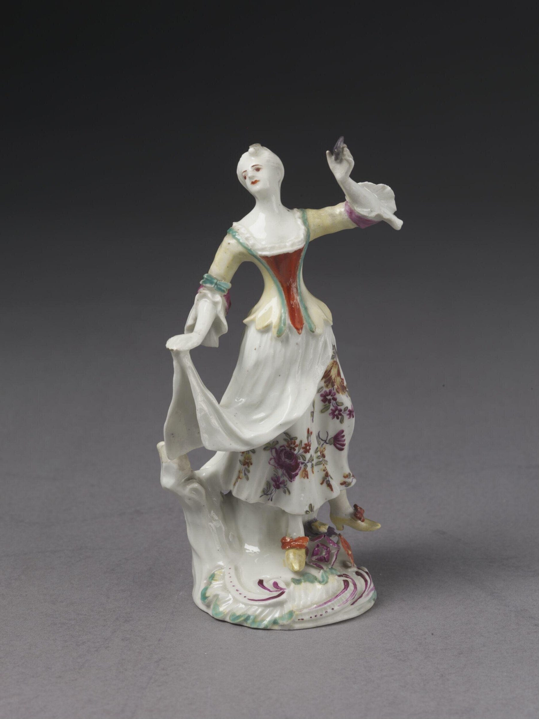 Porcelain figurine of a young woman in red corset and white skirt, left arm raised, right hand gathering her skirt, standing on an ornate base.