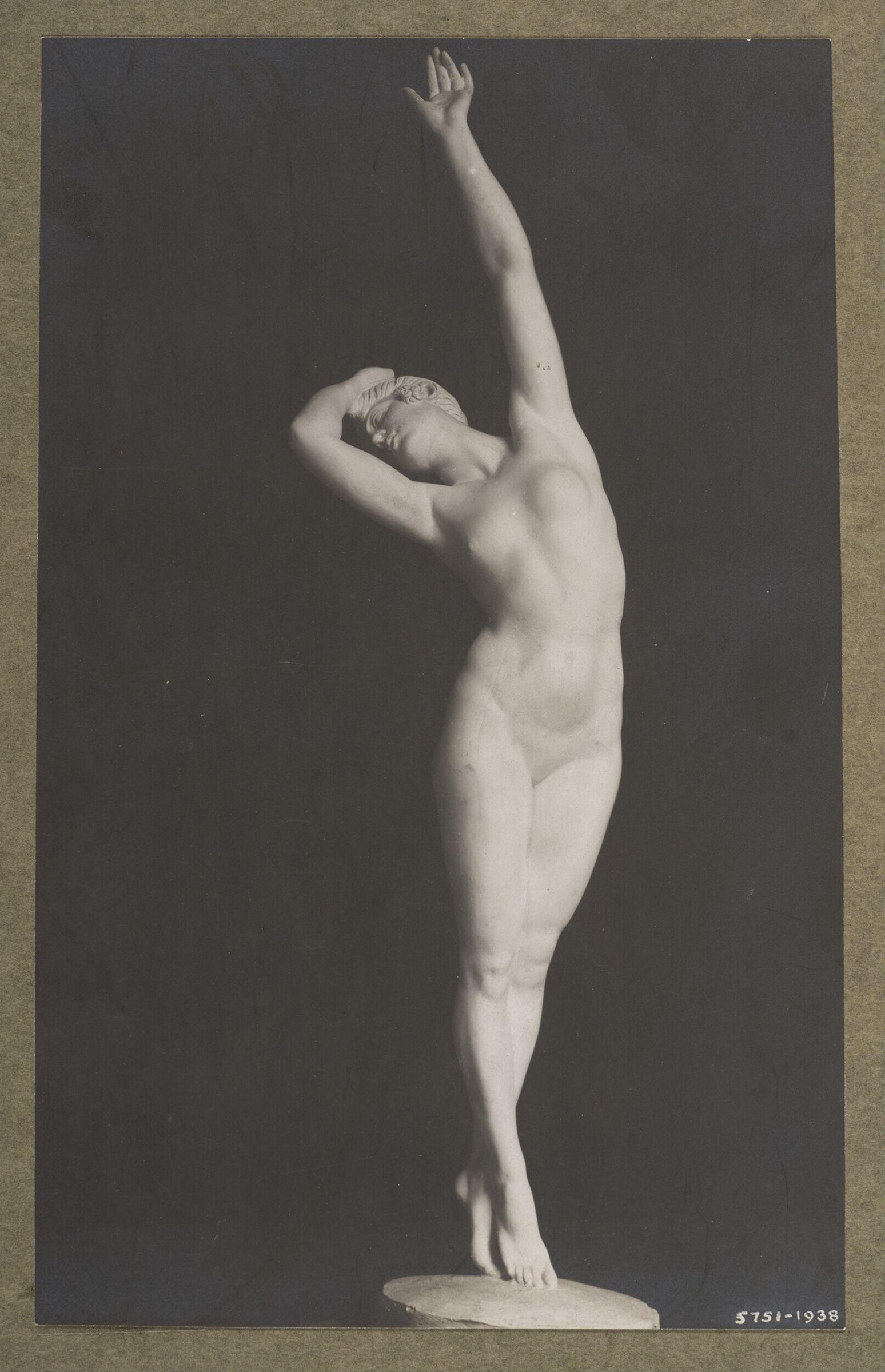 A female dancer stands on one leg against a dark background, her raised arm extended upward, head tilted back, in a classical ballet pose.