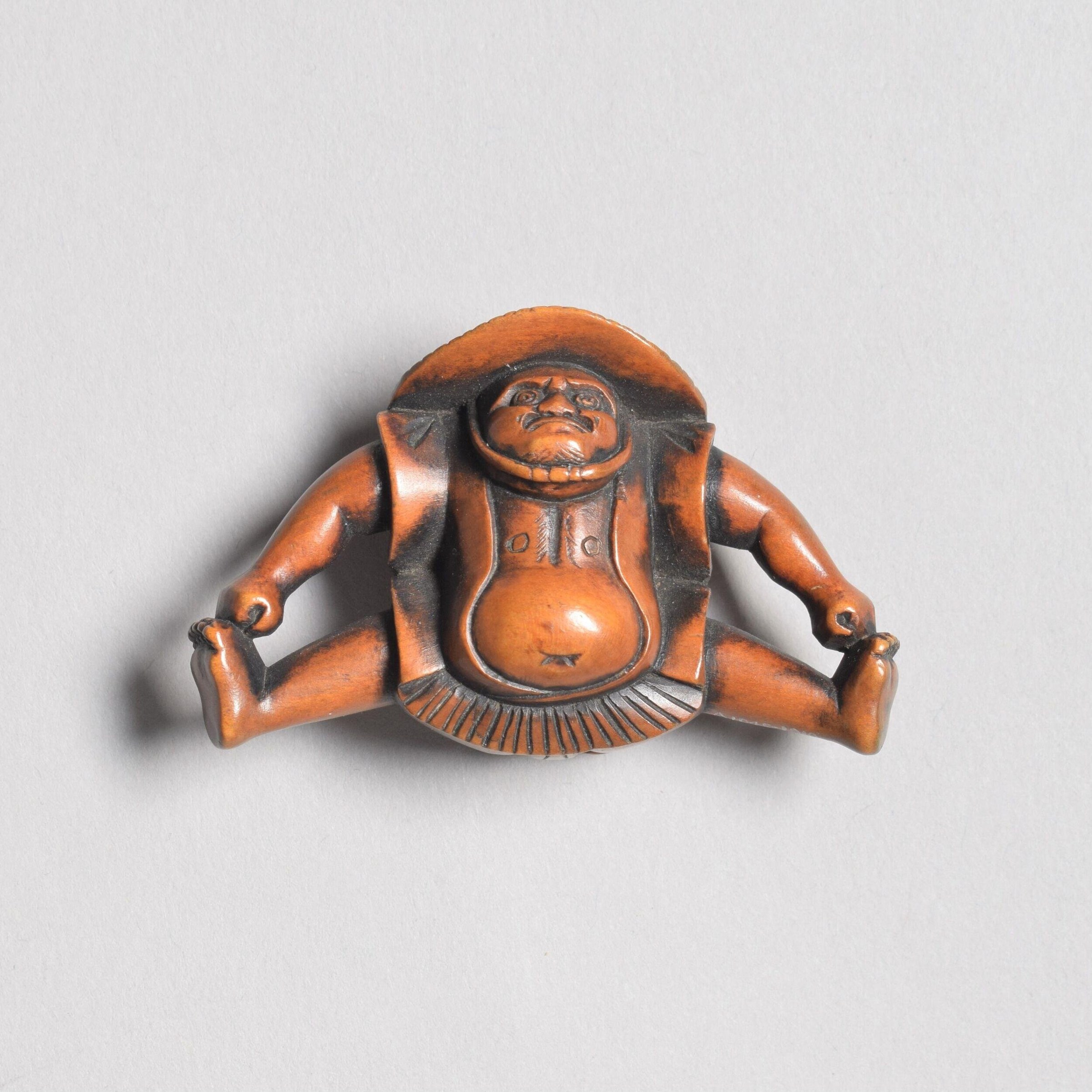 Small wooden carved figure seated with legs splayed, wearing draped robe, with expressive face and rounded belly.