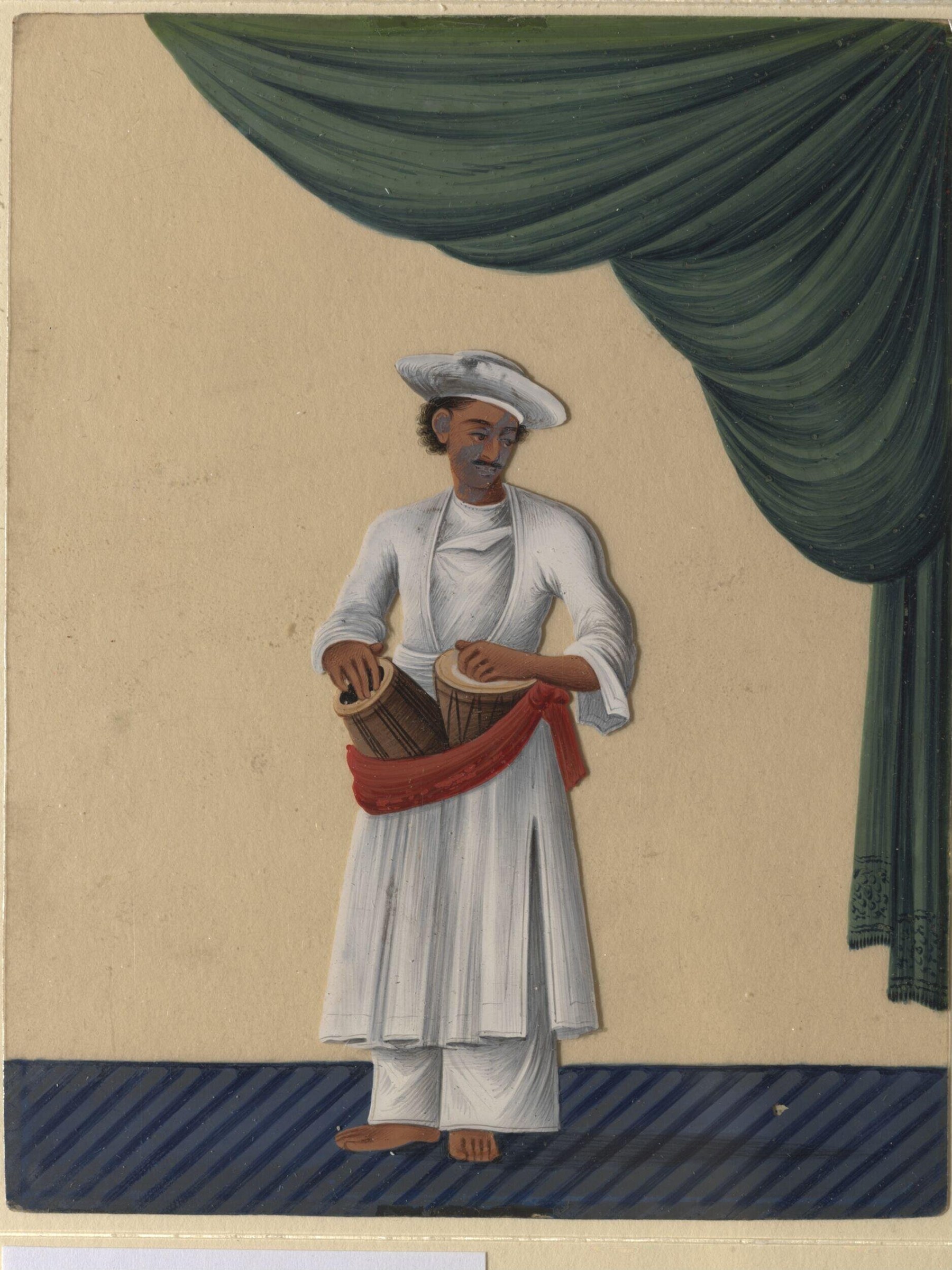 A South Asian man in white kurta and dhoti holds a drum, standing formally against a cream wall with a green draped fabric above.