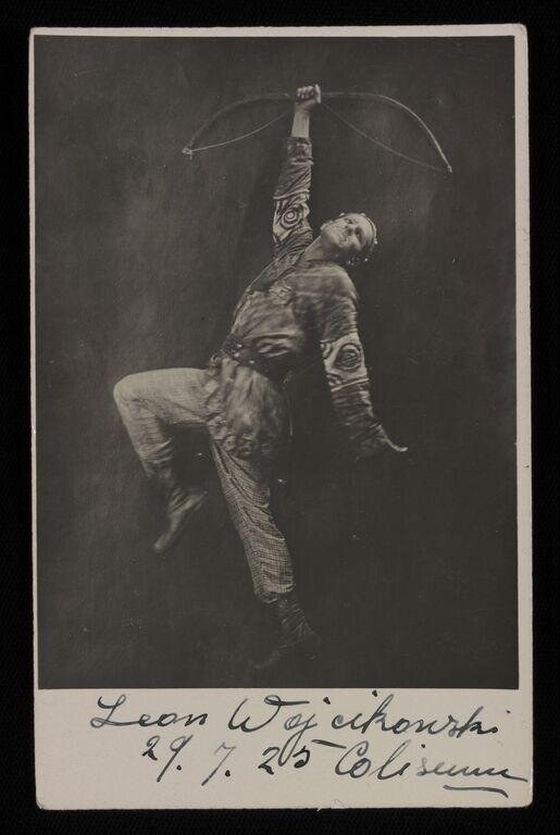 Male dancer suspended in mid-leap, arms raised, body covered in geometric white patterns, wearing light-colored trousers, against dark background