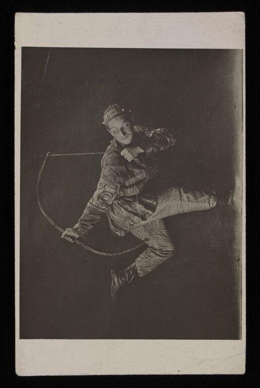 Male ballet dancer in mid-leap, legs split wide, wearing ornamental tunic and headdress, against dark background.
