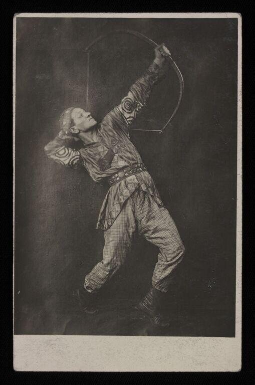 Male dancer in mid-leap, arched backward, holding curved prop aloft, in patterned tunic and leggings against dark background