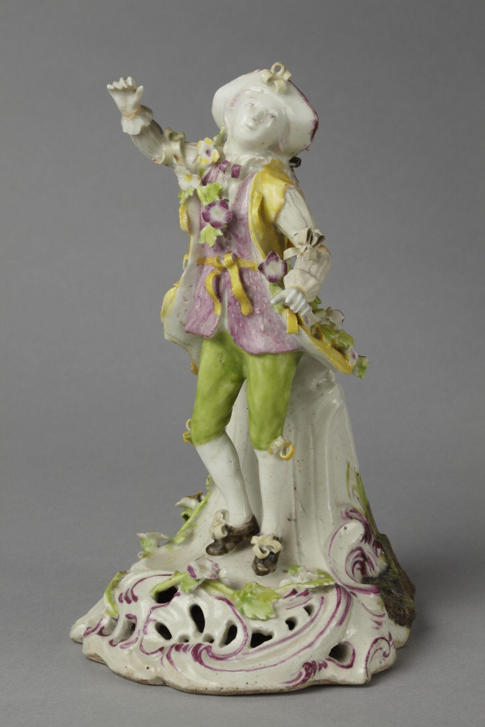 Porcelain figurine of a young man in yellow jacket and green breeches, one arm raised, dancing on a rococo base.