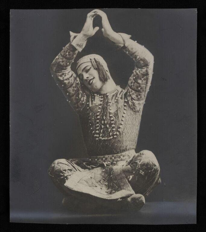 A dancer in an ornately embroidered tunic sits cross-legged with both arms raised above the head, hands meeting in a curved frame, gazing downward.