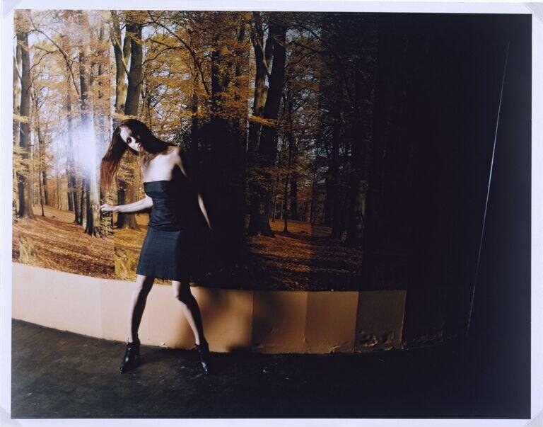 Woman in black strapless dress dances alone against large forest photograph backdrop in gallery space.