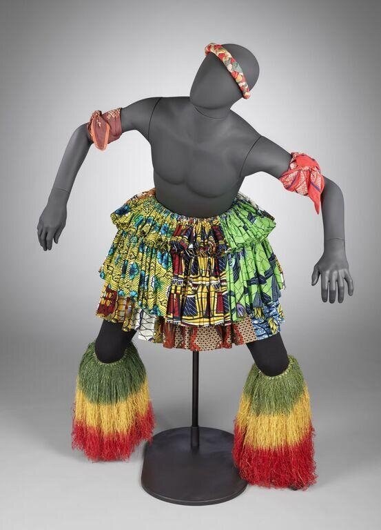 Mannequin wearing vibrant patchwork African textile skirt and dyed raffia leg wrappings in green, yellow, and red, arms spread wide in celebratory gesture.