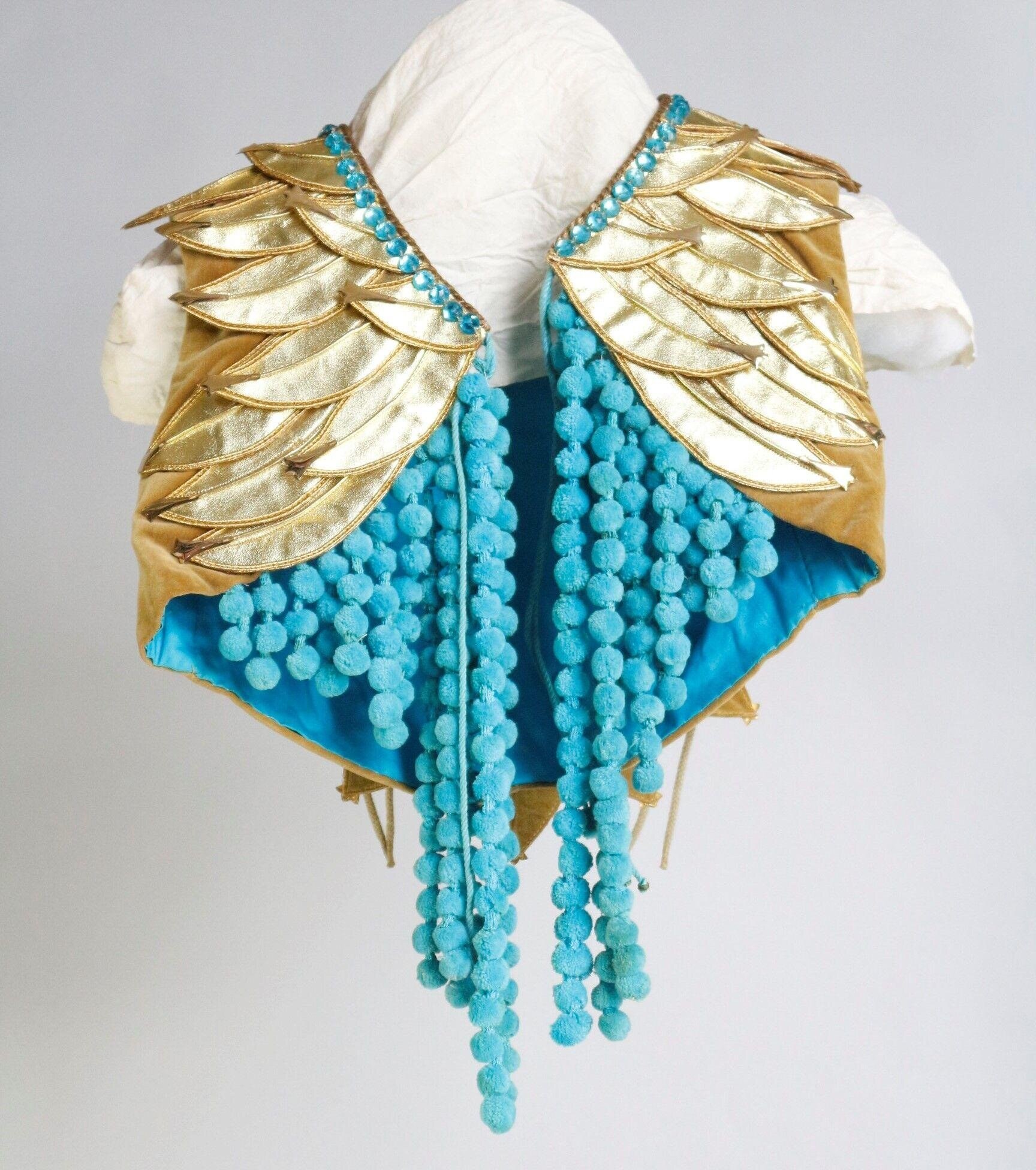 Gold leaf-shaped shoulder mantle with turquoise pom-pom fringe and white wrapped head form, a mid-century theatrical ballet costume component.