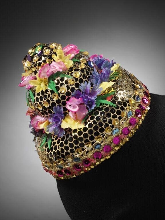 Gold mesh headdress encrusted with layered silk flowers in pink, purple, and yellow, and jeweled beads, displayed on a dress form against dark background.