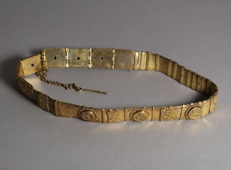 A gilded metal belt with articulated rectangular and circular plaques decorated in repoussé, with a chain attachment, photographed flat against a gray