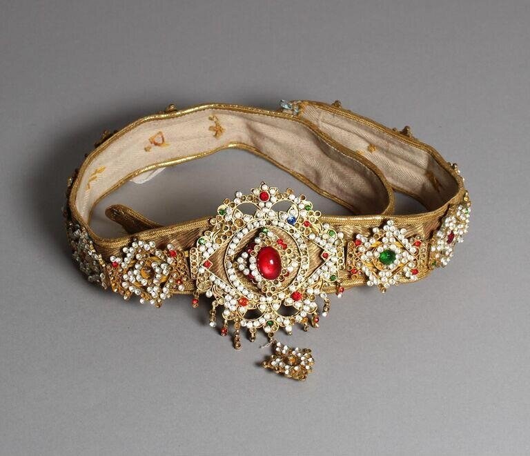 Jeweled gold and silk ornamental belt with central ruby medallion, surrounded by pearls and gemstones, displayed flat.