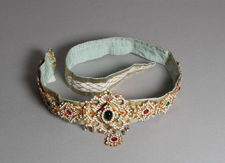Jeweled waistband ornament with pale green pleated silk ribbon and large emerald stone surrounded by seed pearls and gemstones in gold metalwork.