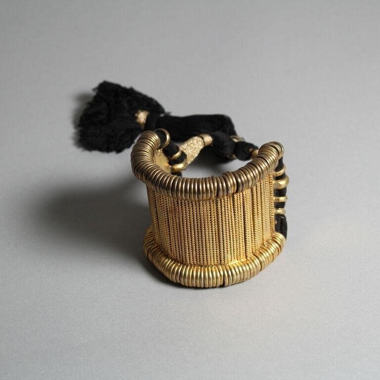 Gold cylindrical cuff with parallel wire construction, coiled trim, and black tassel, photographed against grey background.