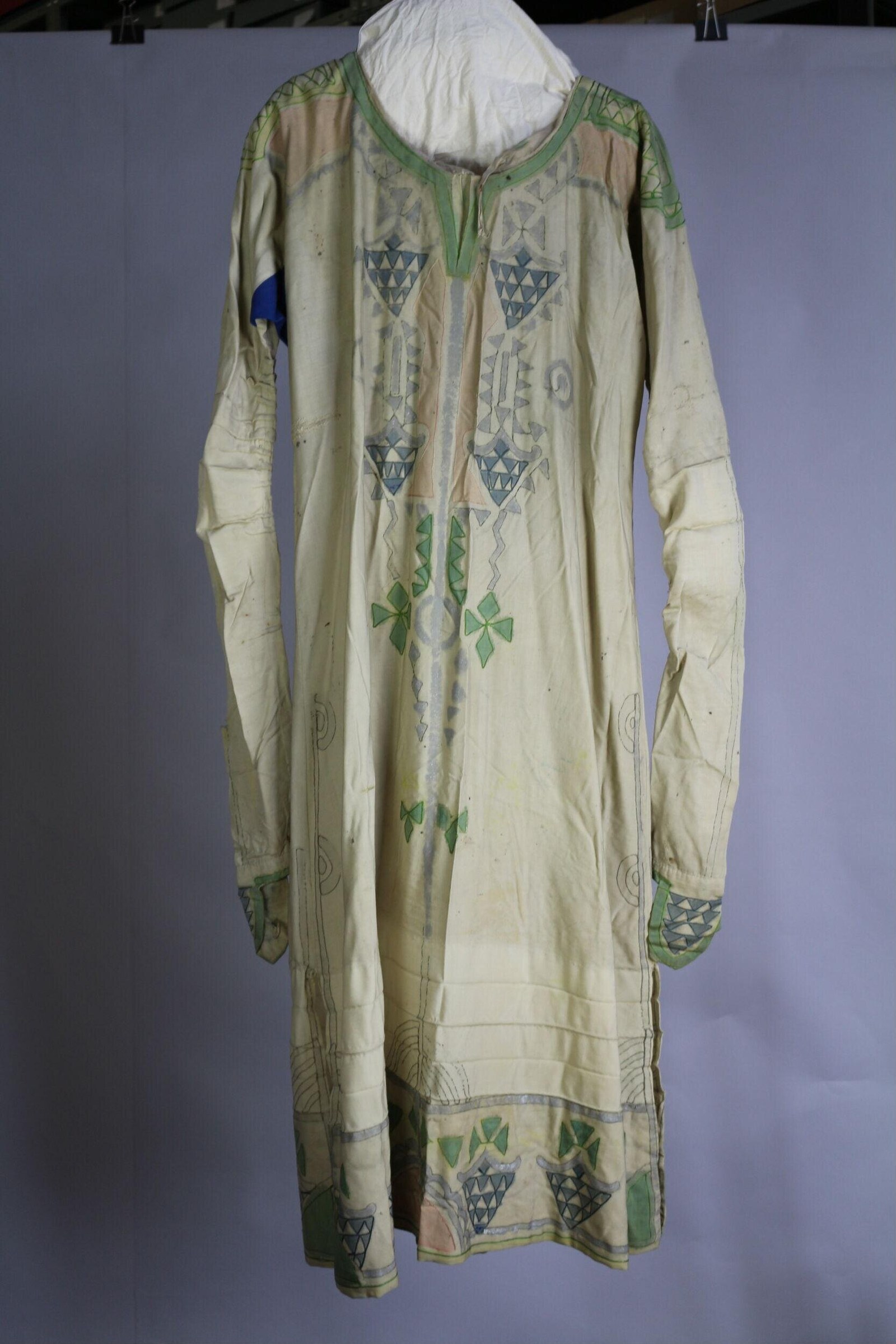 Pale cream linen dress with green trim and blue-grey geometric embroidery on front bodice, sleeves, and hem, displayed on a form.