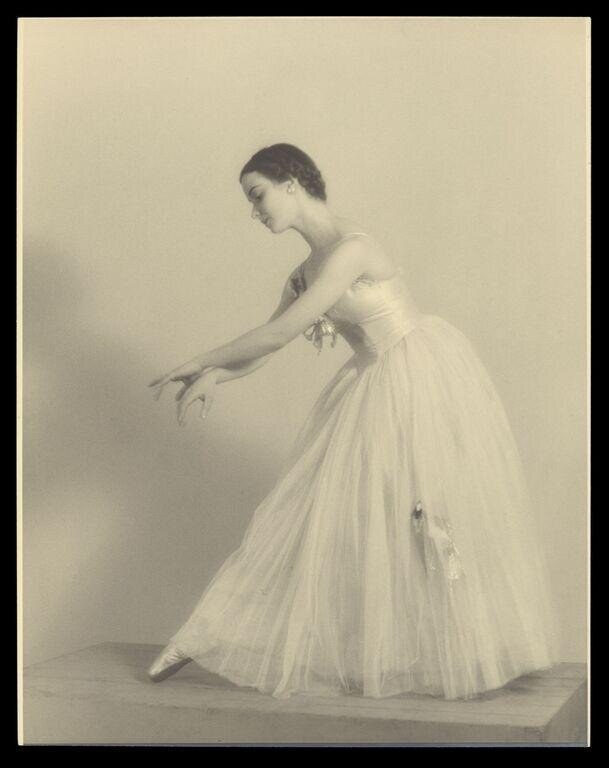 Young female ballet dancer in white tulle tutu and pointe shoes, standing in gentle arabesque, left arm extended, head lowered, against plain background.