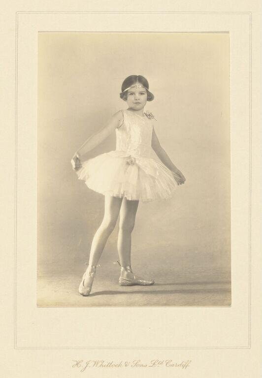 Young girl in white ballet tutu and pointe shoes stands in calm pose against plain background in studio portrait.