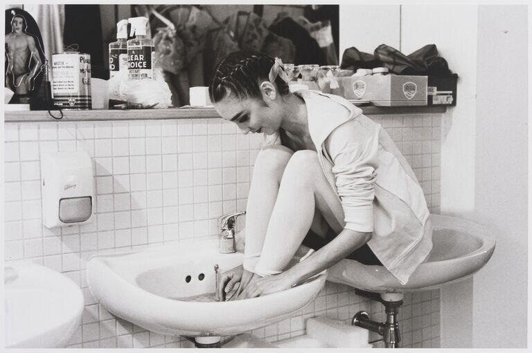 A woman in a white sleeveless shift crouches inside a white porcelain bathroom sink, her body compressed and folded.