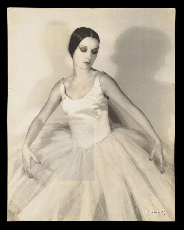 Studio portrait of a ballet dancer in a white leotard and tulle skirt, gazing downward, circa 1930.