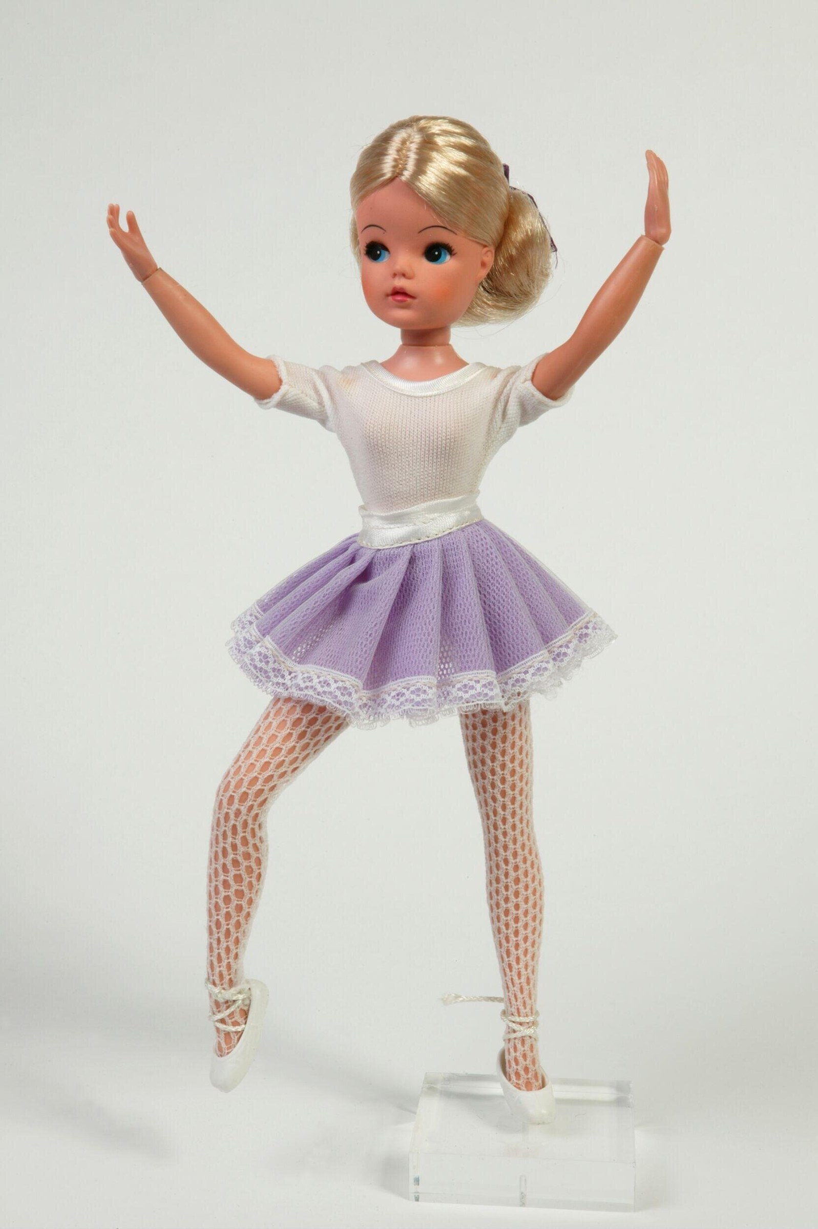 A blonde doll in a white shirt and lavender tulle skirt with white pointe shoes stands with arms raised, smiling.