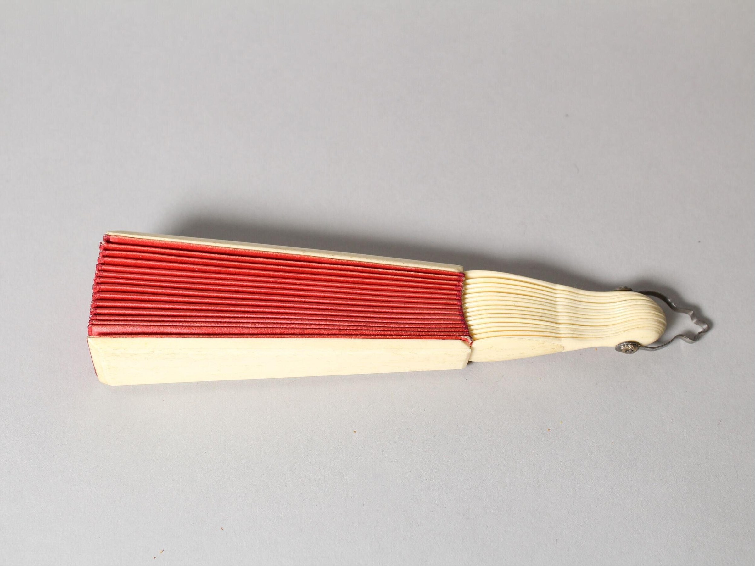 Red pleated silk fan with ivory bone handle, laid flat and fully open, casting shadow on neutral background.