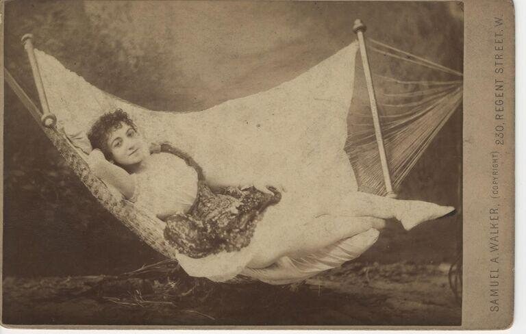 Woman in patterned dress reclines in draped swing, studio photograph on cardboard mount, 1885.