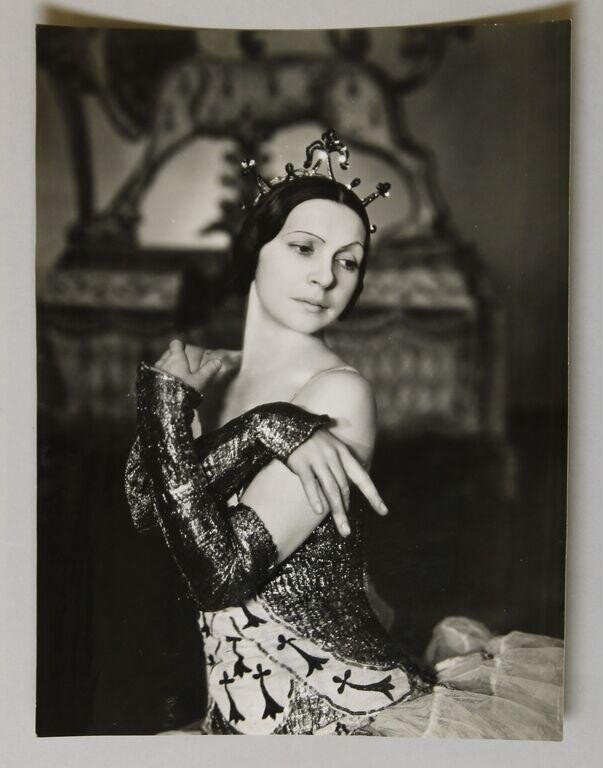 A female ballet dancer in a black-and-white studio photograph wears an ornate off-shoulder tunic, patterned skirt, and tiara, posed in classical ballet stance.
