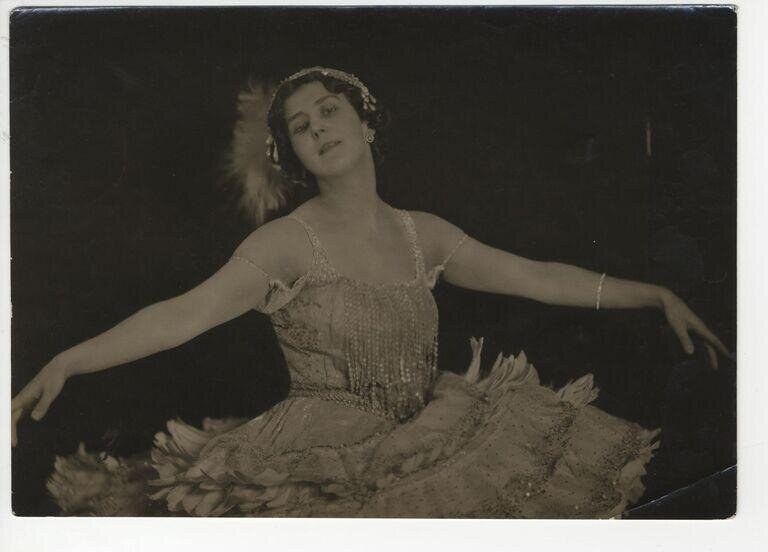 A female ballet dancer in a beaded tunic holds an arabesque pose, one leg extended behind, against a black background.