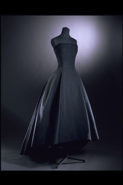 A dark voluminous evening dress on a headless dress form, lit dramatically from above against a dark background.