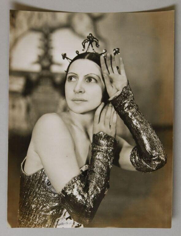 Woman in sequined costume and ornate headdress gazes upward with hand raised to her face in a moment of reverie.