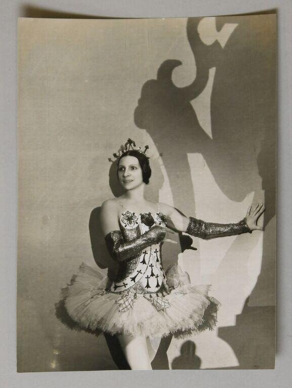 Female ballet dancer in jeweled bodice and white tulle tutu with tiara stands formally against a theatrical backdrop, 1935.