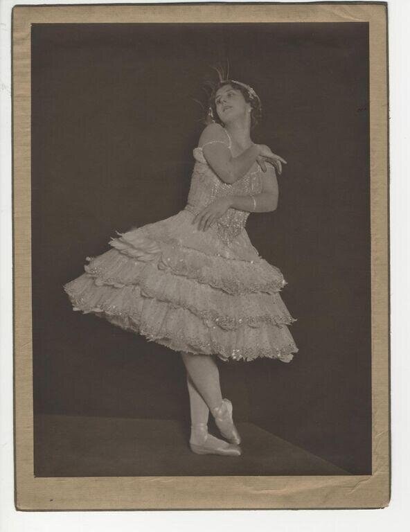 A young woman in a white layered lace dress stands in a classical ballet pose against a dark background, weight on one leg, the other extended with pointed