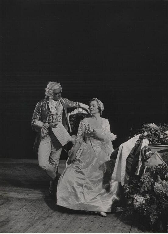A man in 18th-century coat and breeches holds sheet music while dancing with a woman in a pale gown on a dark stage.