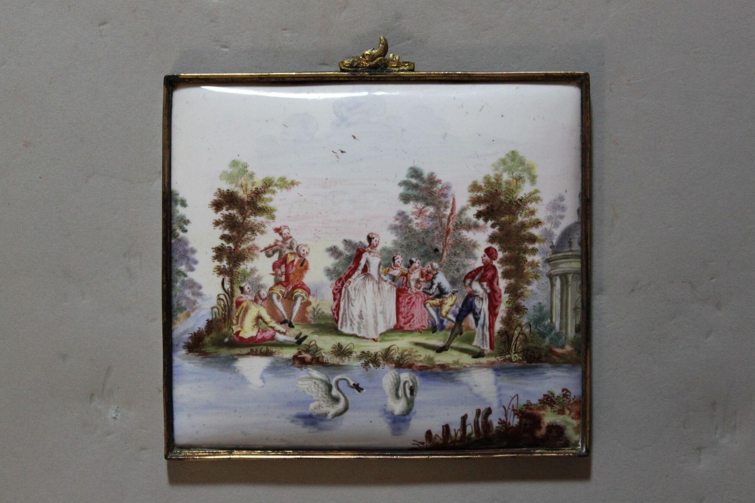 Hand-painted porcelain plaque showing elegantly dressed 18th-century figures in a garden beside water, with swans and trees framing the scene.