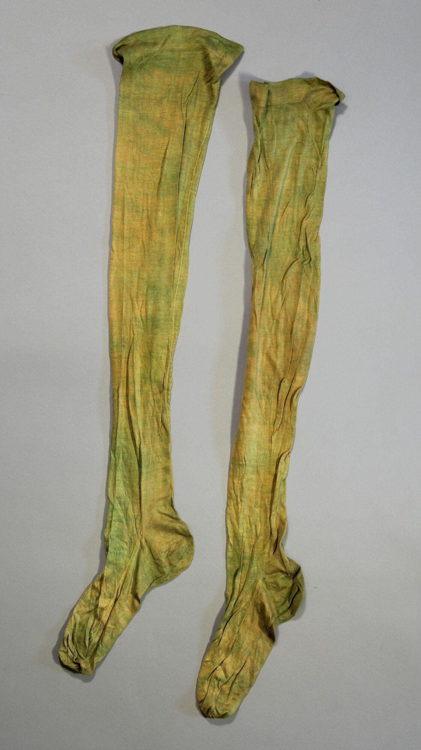Two pale yellow-green linen stockings laid flat, wrinkled and creased, with visible seams and a shaped foot.