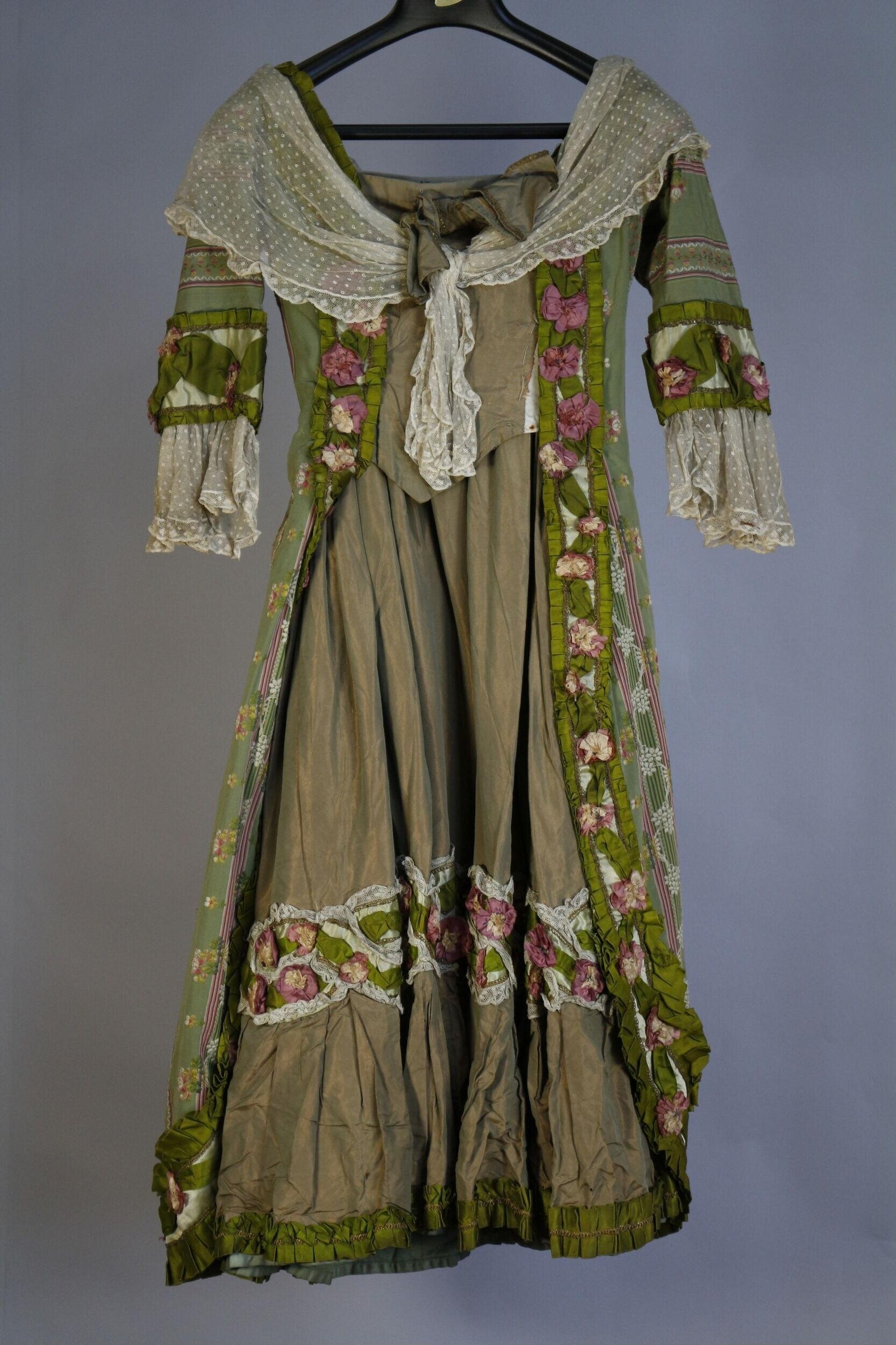 Early 1910s theatre costume: cream linen dress with sage green floral panels and tiered skirt, displayed on a hanger.
