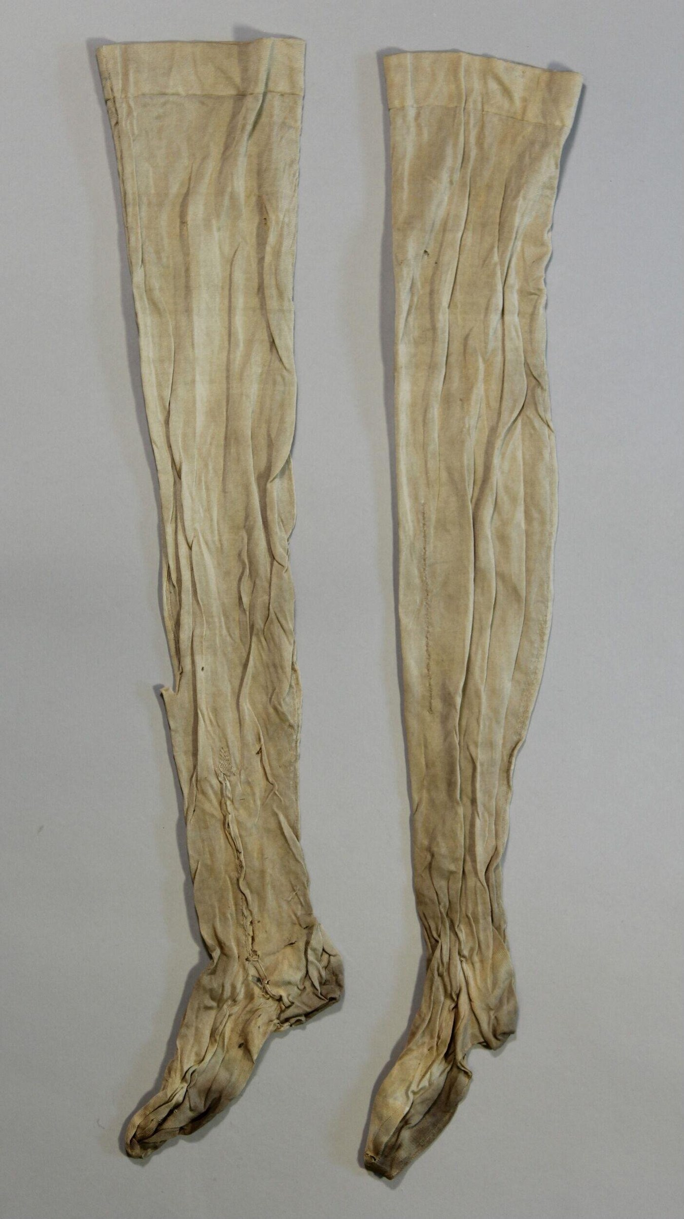 Pair of cream-coloured linen stockings with pronounced vertical creasing and bunched fabric at the feet, laid out vertically.