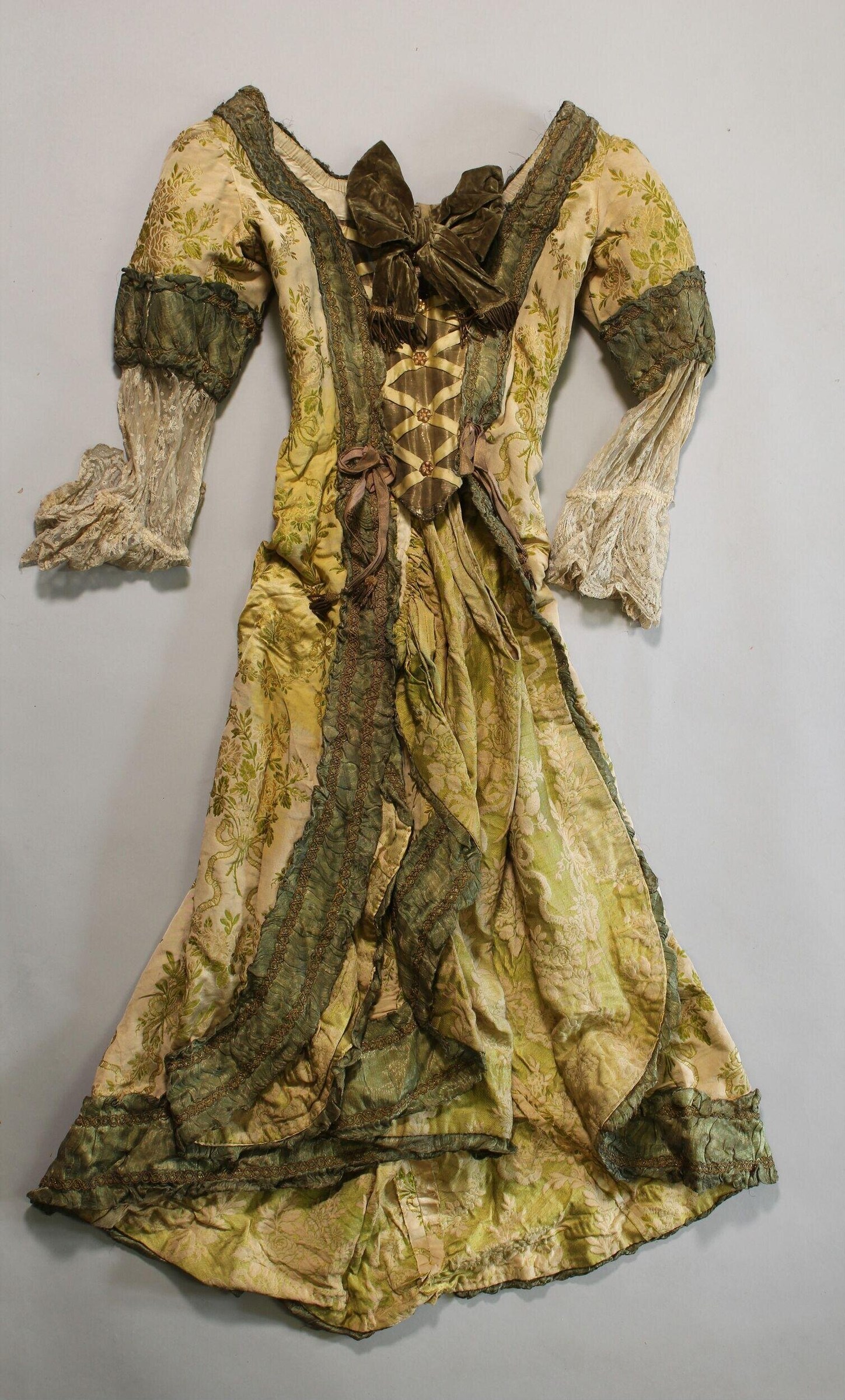Full-length Edwardian theatre dress in cream silk with deep green velvet trim, displayed flat, showing voluminous draped sleeves and cinched waist.