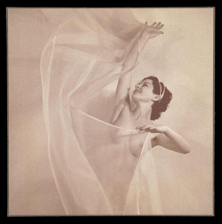 Female dancer in pale draped dress, arching backward with left arm extended upward, billowing translucent fabric swirling around her upper body.